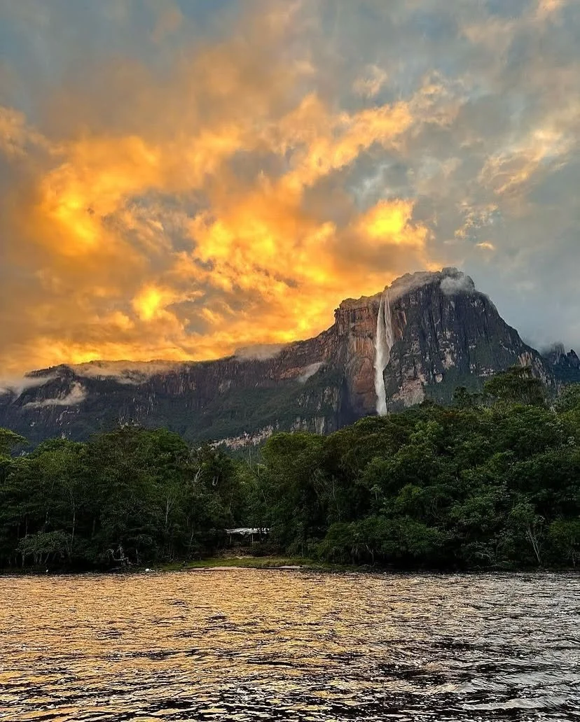 Sunset over a mountain with a waterfall, surrounded by trees, with a river in the foreground.