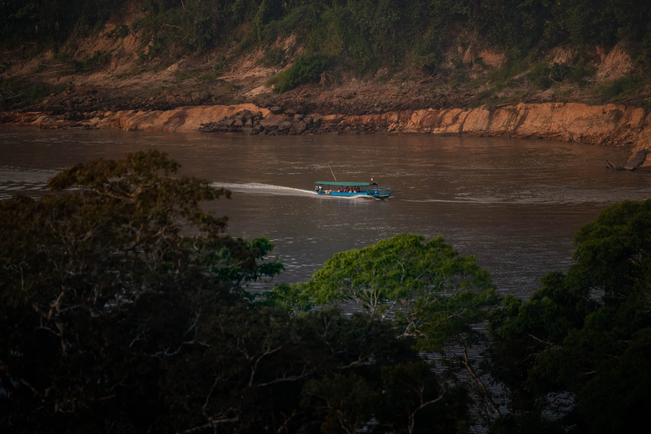 A blue boat with people on board is moving across a river surrounded by green bushes and rocky brown cliffs.