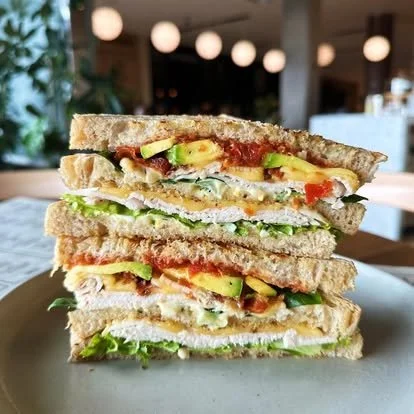 Stacked sandwiches with avocado, tomato, lettuce, and chicken on toasted bread in a restaurant setting.
