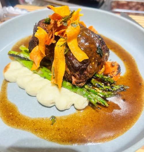 A plated dish with beef rolled and topped with crispy vegetables, served with mashed potatoes and asparagus, in a brown gravy sauce.