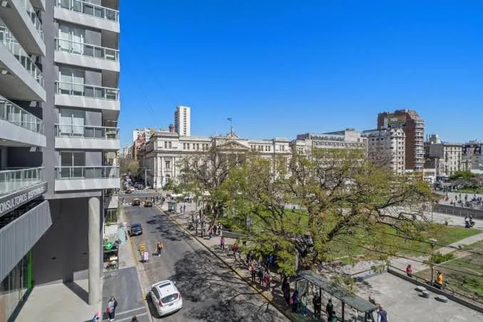 A city street scene with a tree-lined sidewalk, pedestrians, cars, and a historic building under a clear blue sky.