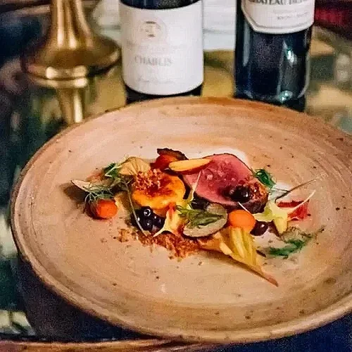 A wooden plate with a small portion of gourmet food, including a piece of cooked meat, colorful vegetables, and garnishes, with wine bottles in the background.
