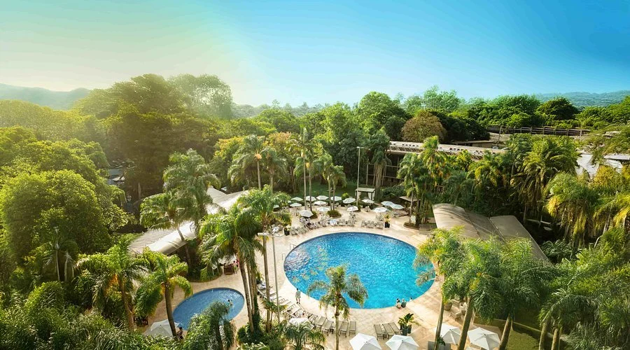 Aerial view of a lush tropical resort with a large swimming pool, smaller pool, numerous lounge chairs with umbrellas, surrounded by palm trees and greenery, and distant hills in the background.