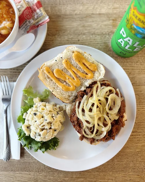 Plate with a sandwich featuring mustard, a scoop of macaroni salad with black olives on lettuce, and a fried beef patty topped with sliced onions.