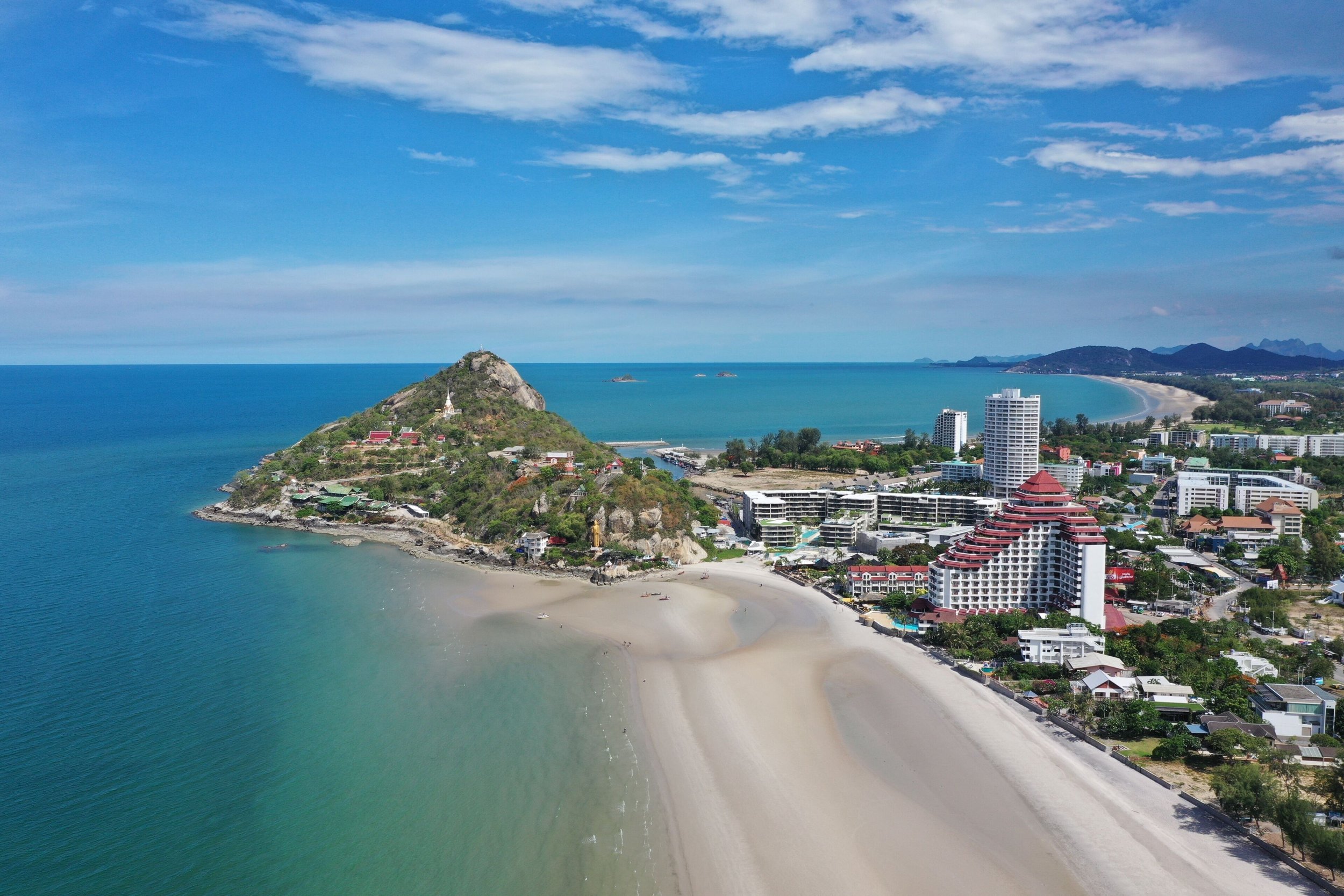 Aerial view of a coastal city with a beach, high-rise hotels, and a hill with a shrine. The ocean is on the left with calm blue water, and the sky is partly cloudy.