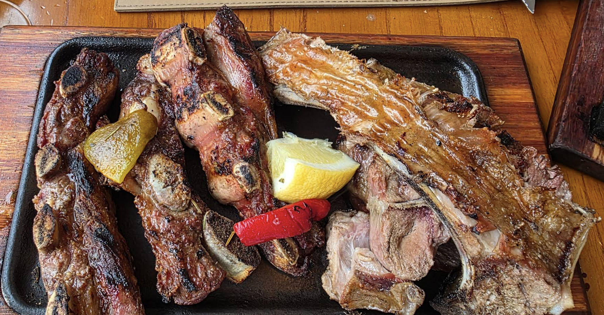 Assorted grilled meats served on a black sizzling platter, garnished with lemon, red chili pepper, and pickled pepper, on a wooden table.