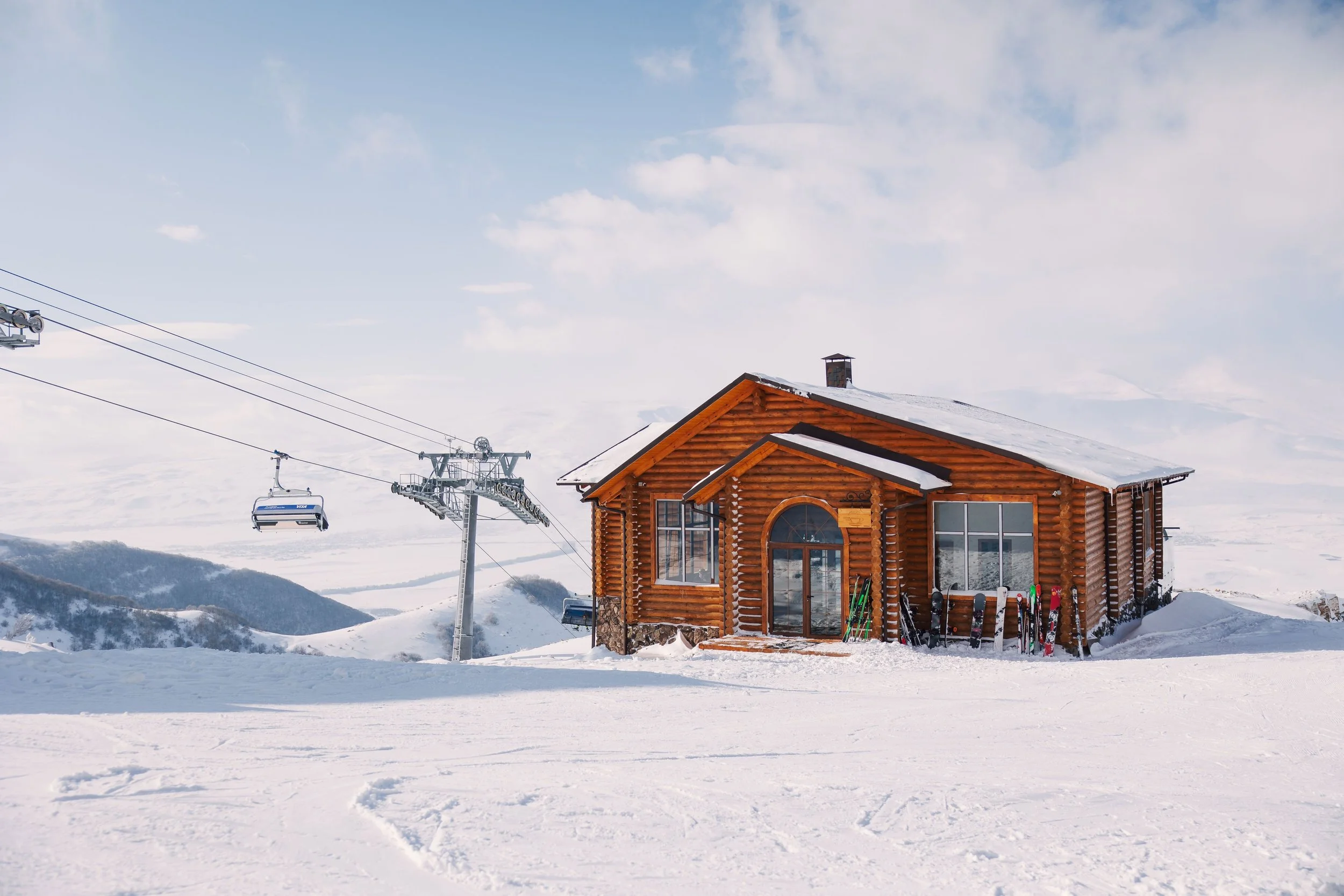 A wooden cabin in a snowy landscape with ski equipment outside and ski lift in the background.