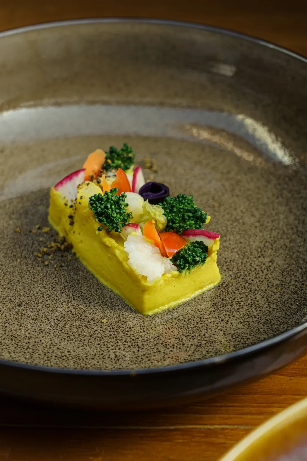A rectangular slice of yellow polenta topped with an assortment of colorful vegetables, including broccoli, radishes, carrots, and cauliflower, served on a ceramic plate.