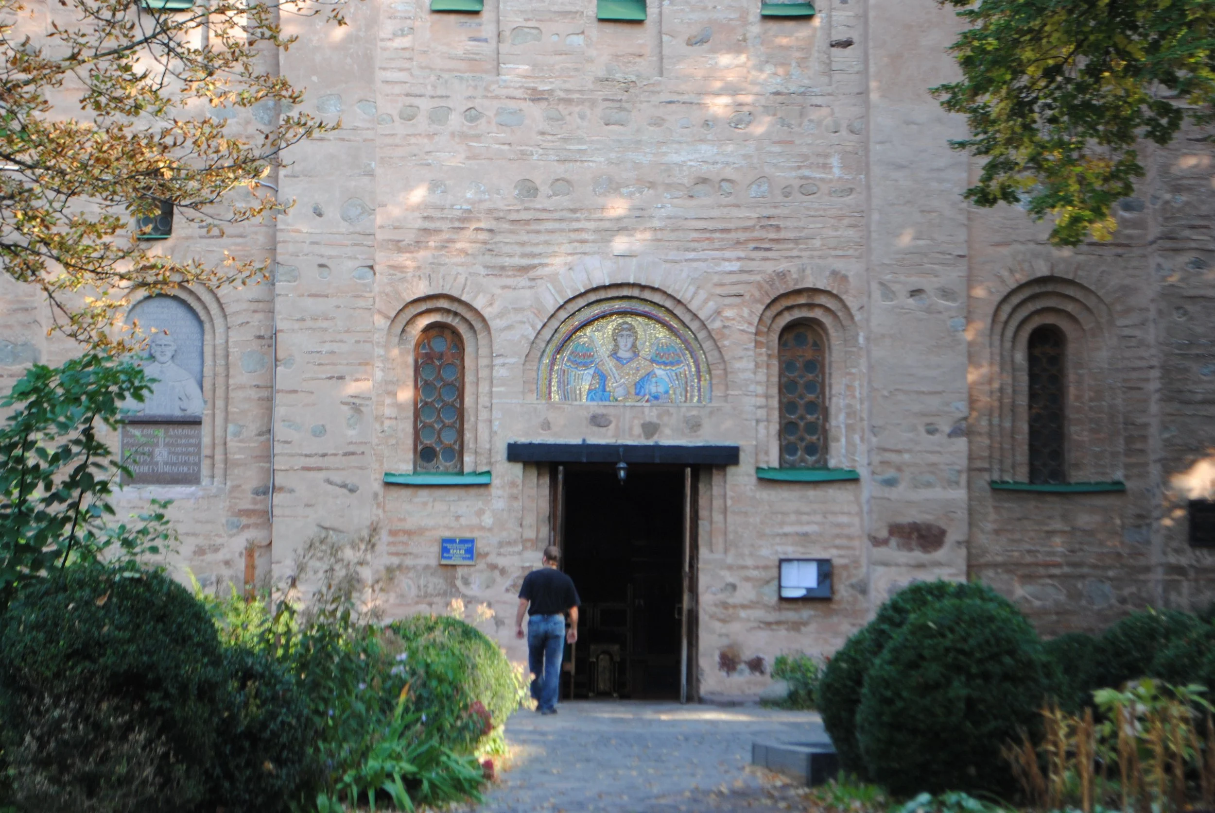 A stone church facade with a mosaic of an angel above the entrance, surrounded by trees and greenery, with two people walking toward the door.