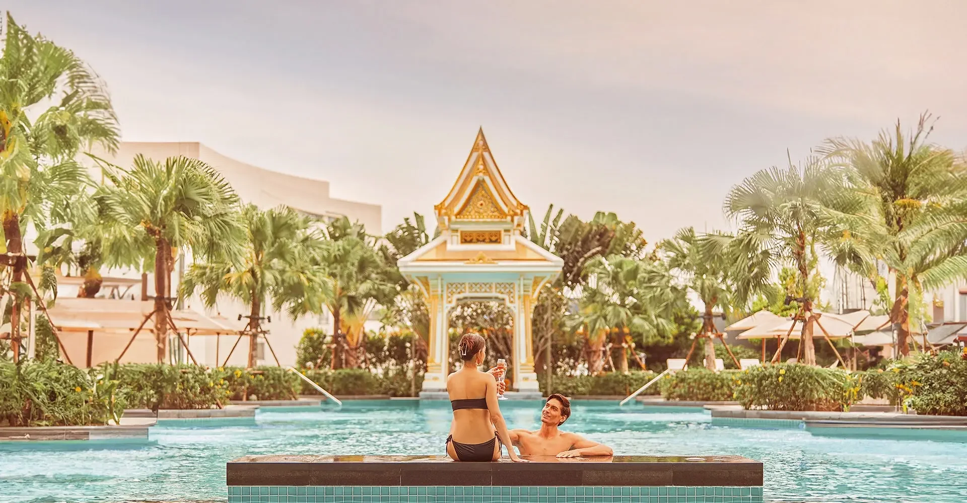 A couple enjoying a relaxing moment in a swimming pool at a luxurious resort with tropical plants and traditional Thai architecture in the background.