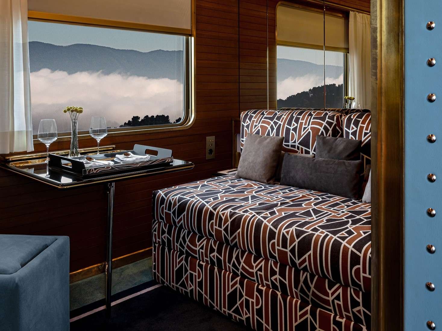 A cozy room with a window overlooking mountains and clouds, a patterned sofa with gray pillows, a small table set with wine glasses and a tray, and warm wooden walls.