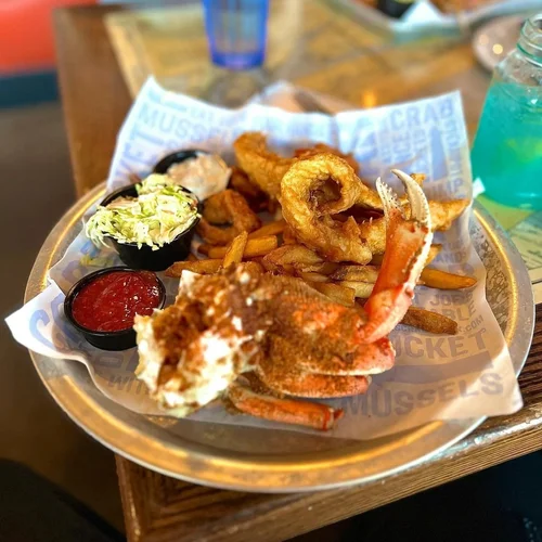 A seafood platter with fried shrimp, onion rings, coleslaw, French fries, and dipping sauces on a metal tray lined with paper.