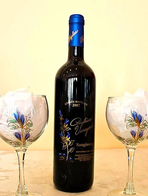 A bottle of red wine with a blue foil cap, flanked by two wine glasses with painted floral designs