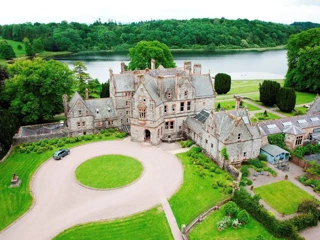 A large historic castle with gray stone walls and towers, surrounded by green lawns and trees, with a circular driveway and a lake in the background.