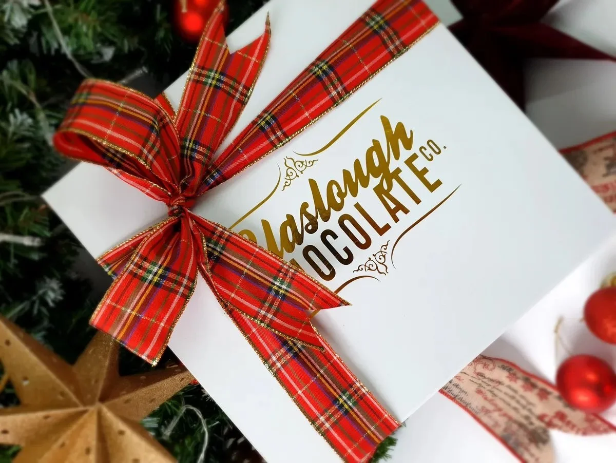 A white gift box with gold and brown logo reading, 'Paslagh Chocolate Co.', adorned with a red plaid ribbon tied in a bow.