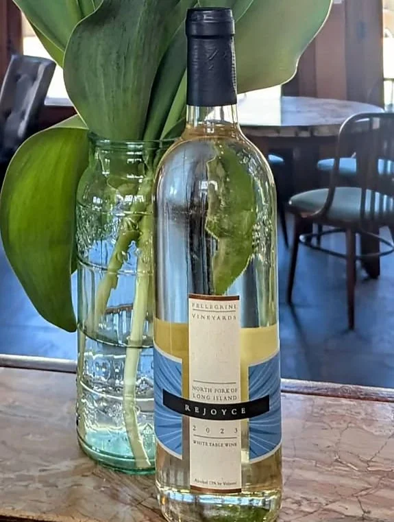 A bottle of wine placed in front of a vase with green leafy plant on a wooden table in a restaurant or cafe setting.