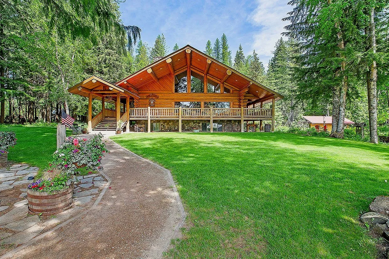 A large wooden house with a mountainous roof, surrounded by tall pine trees and a green lawn. A curved stone pathway leads to the house, which has a balcony with railings and steps on the left side. Flowers in a barrel planter are at the start of the pathway. The sky is partly cloudy.