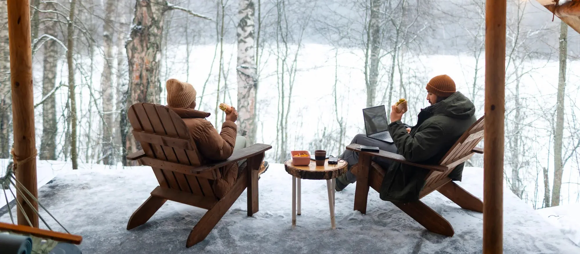 Two people sitting on wooden chairs outside on a snowy deck, enjoying hot drinks and snacks, with a forest in the background.