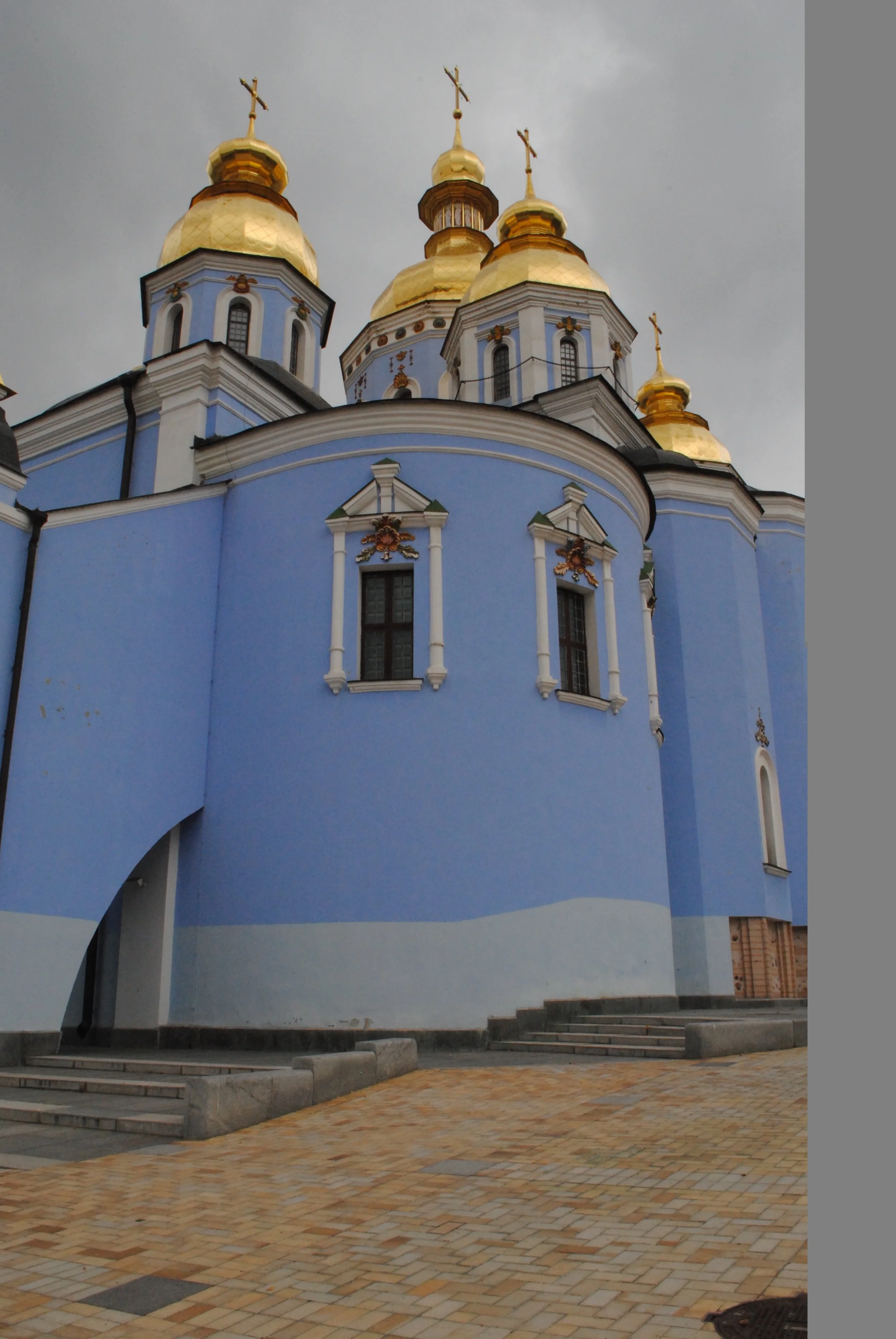 A view of a blue church with five golden domes topped with crosses, set against a cloudy sky, with stone steps and a paved courtyard in the foreground.