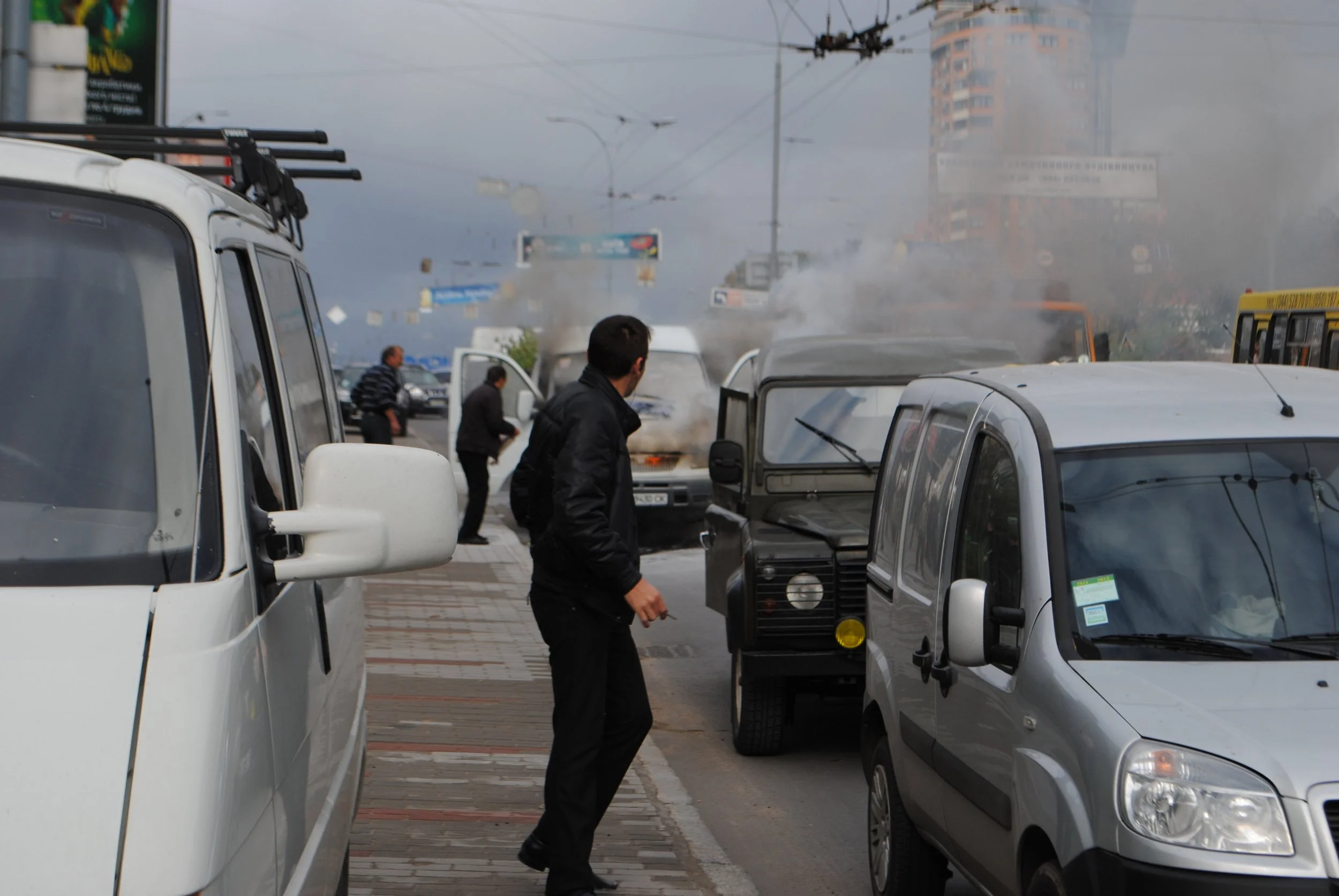 Street scene with multiple parked cars, a person walking, and smoke or fog in the background. Kiev November 2011, Marina Parrish ©