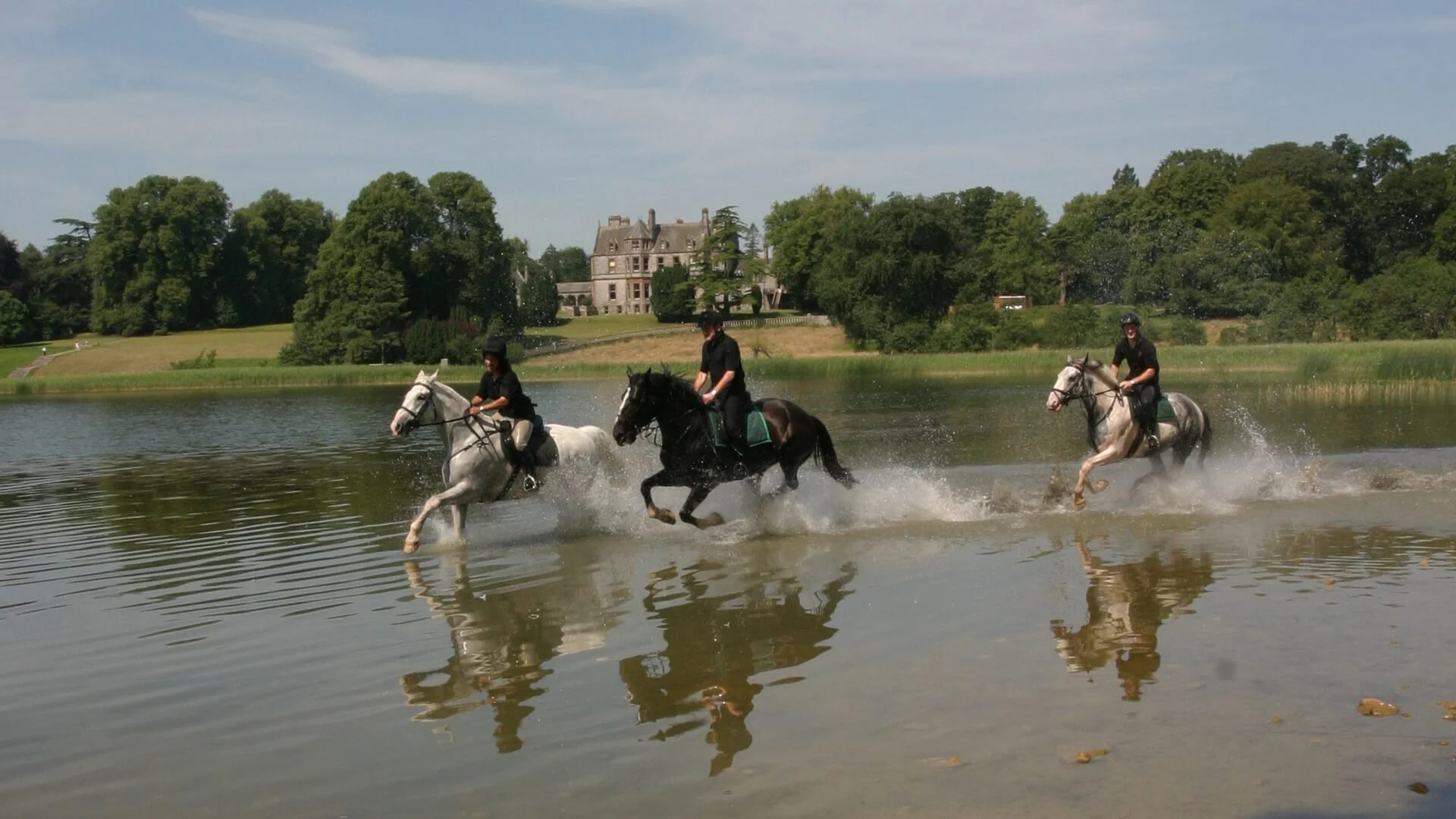 Three horseback riders crossing a lake with splashes of water, in front of a large estate surrounded by trees under a partly cloudy sky.