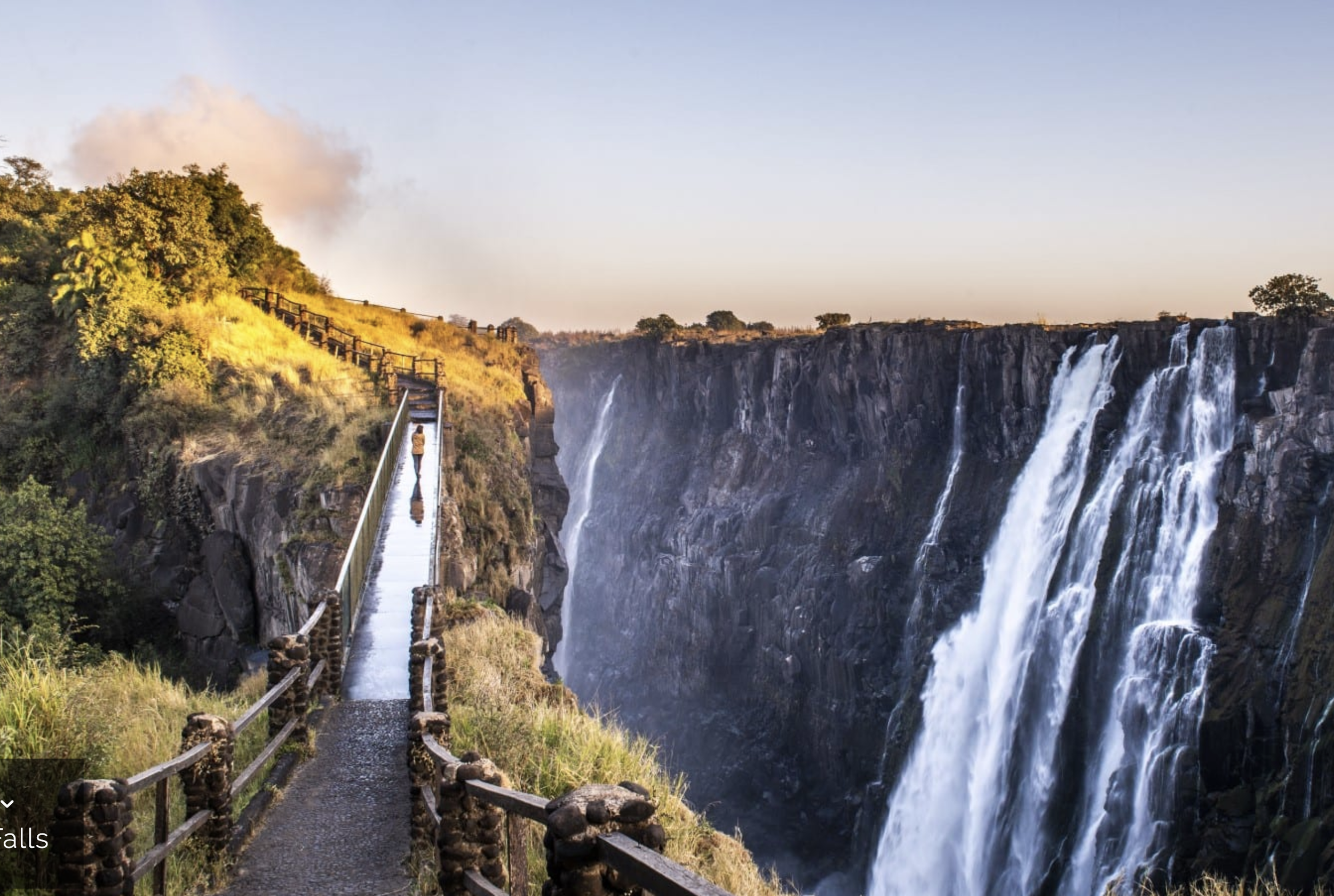 A walkway with wooden railings along a cliff overlooking a waterfall with cascading water, green foliage, and a partly cloudy sky.