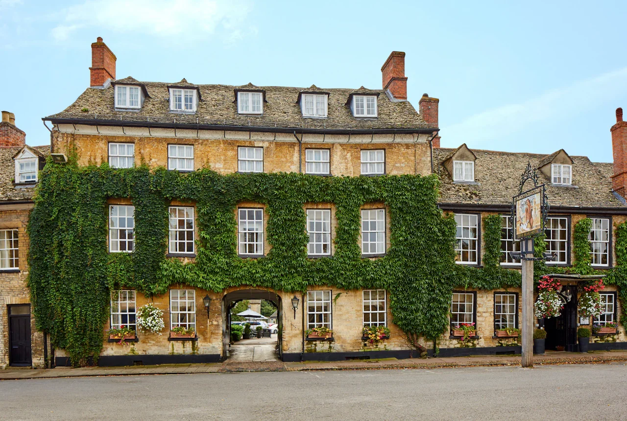 A historic building covered partly with green climbing ivy, with multiple windows, flower boxes, and a sign hanging outside that appears to be a pub or restaurant.