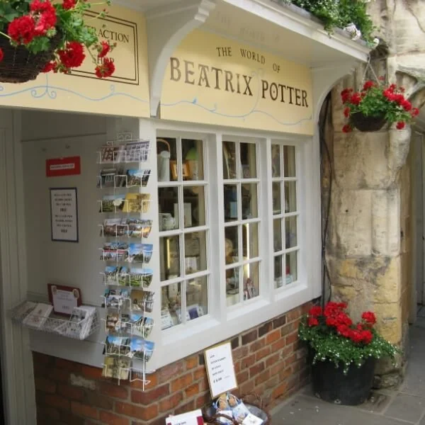 A bookstore storefront with a sign reading "The World of Beatrix Potter" and a display window. There are red flowers in hanging baskets and a large potted plant with red flowers on the sidewalk.