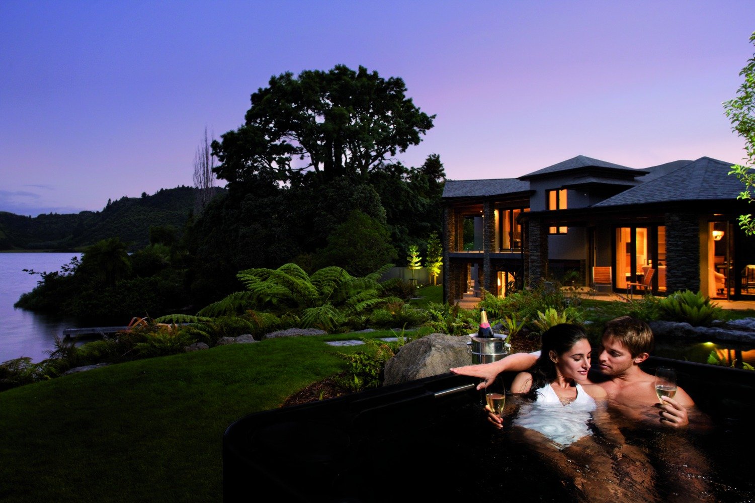 A couple enjoying a hot tub outdoors at dusk, holding glasses of wine, with a modern house and lush landscape in the background.