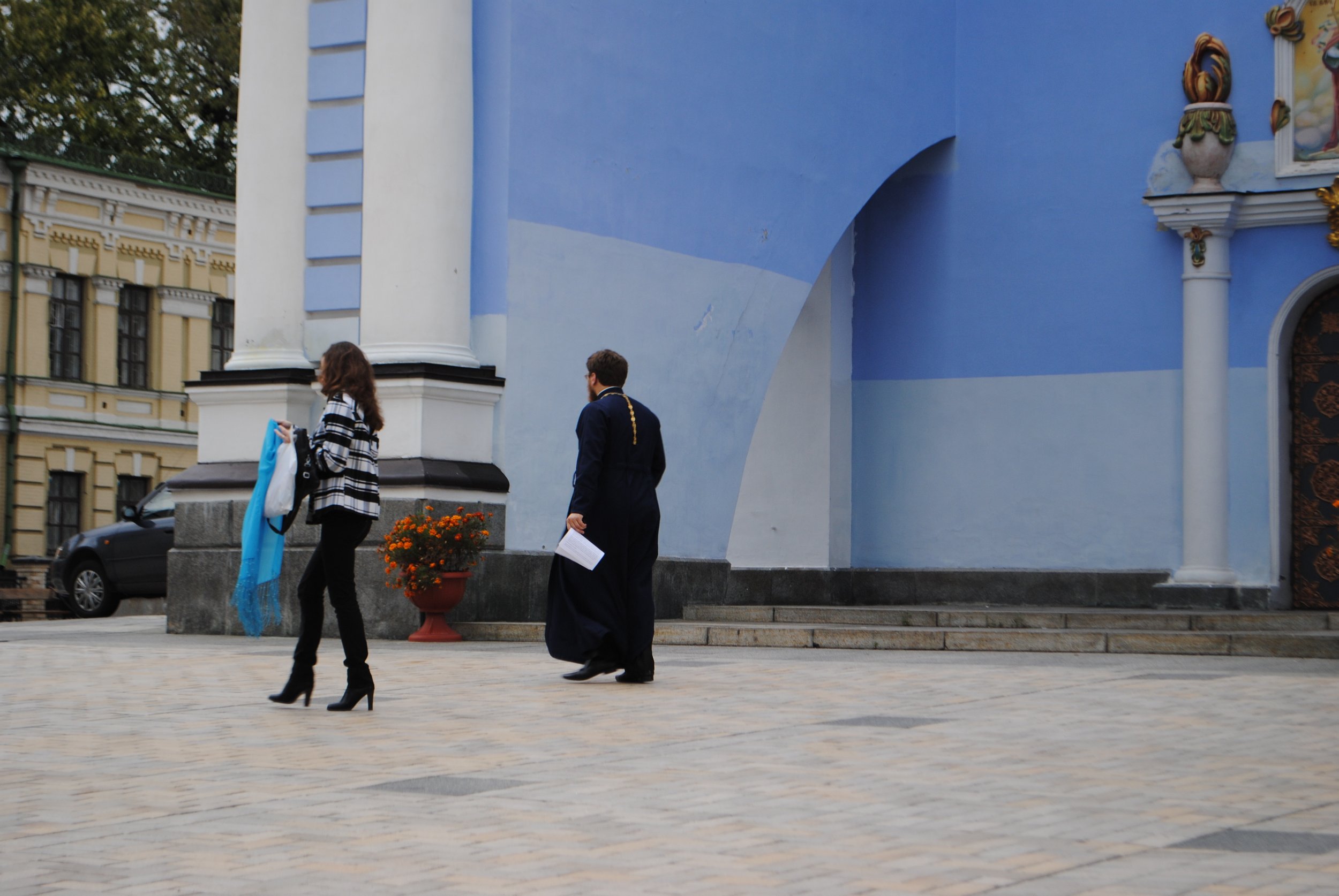 Two women and one man walking outside near a blue building with ornate decorations. The women are dressed in stylish outfits with high heels, and the man is dressed formally carrying papers.