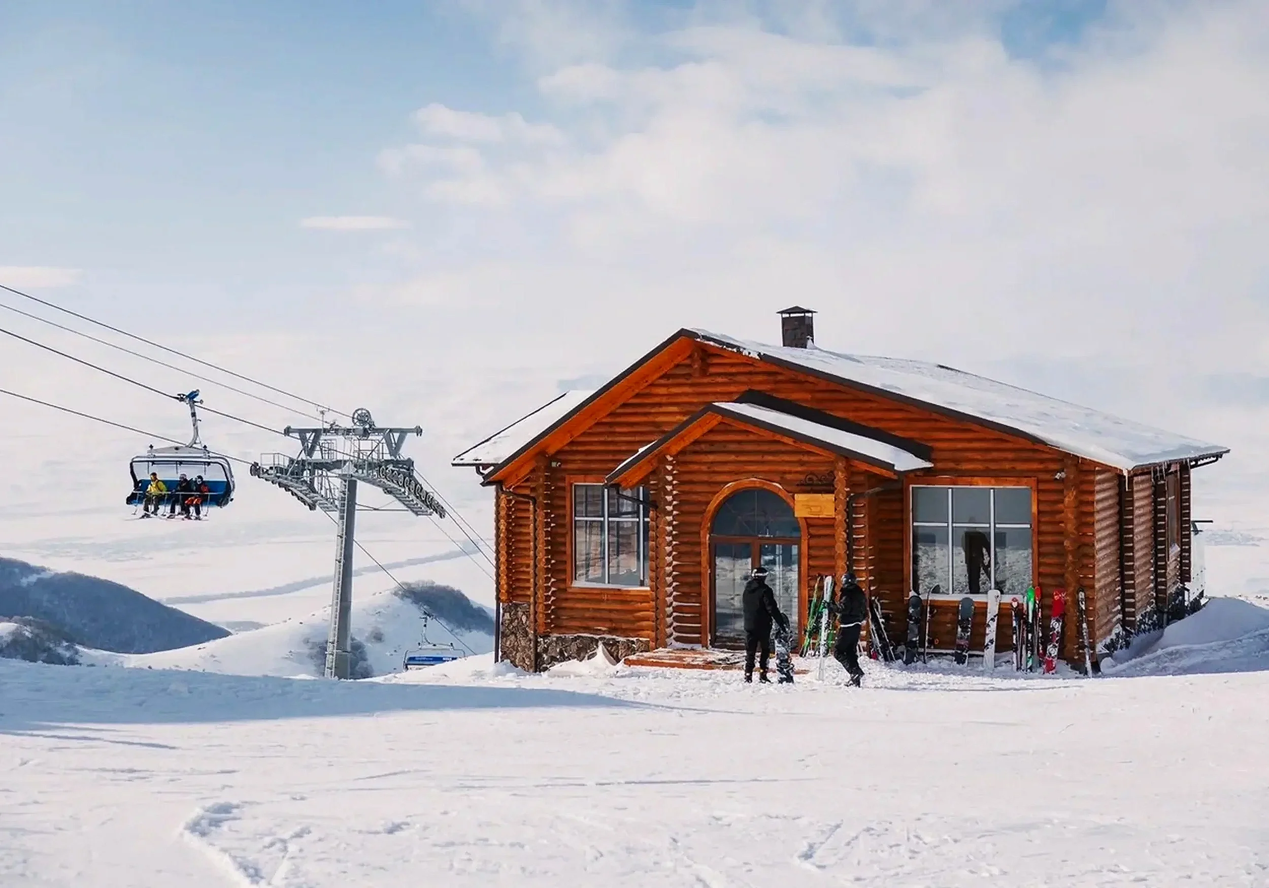 A wooden vacation chalet on a snowy mountain with a ski lift carrying skiers in the background and snow-covered landscape.