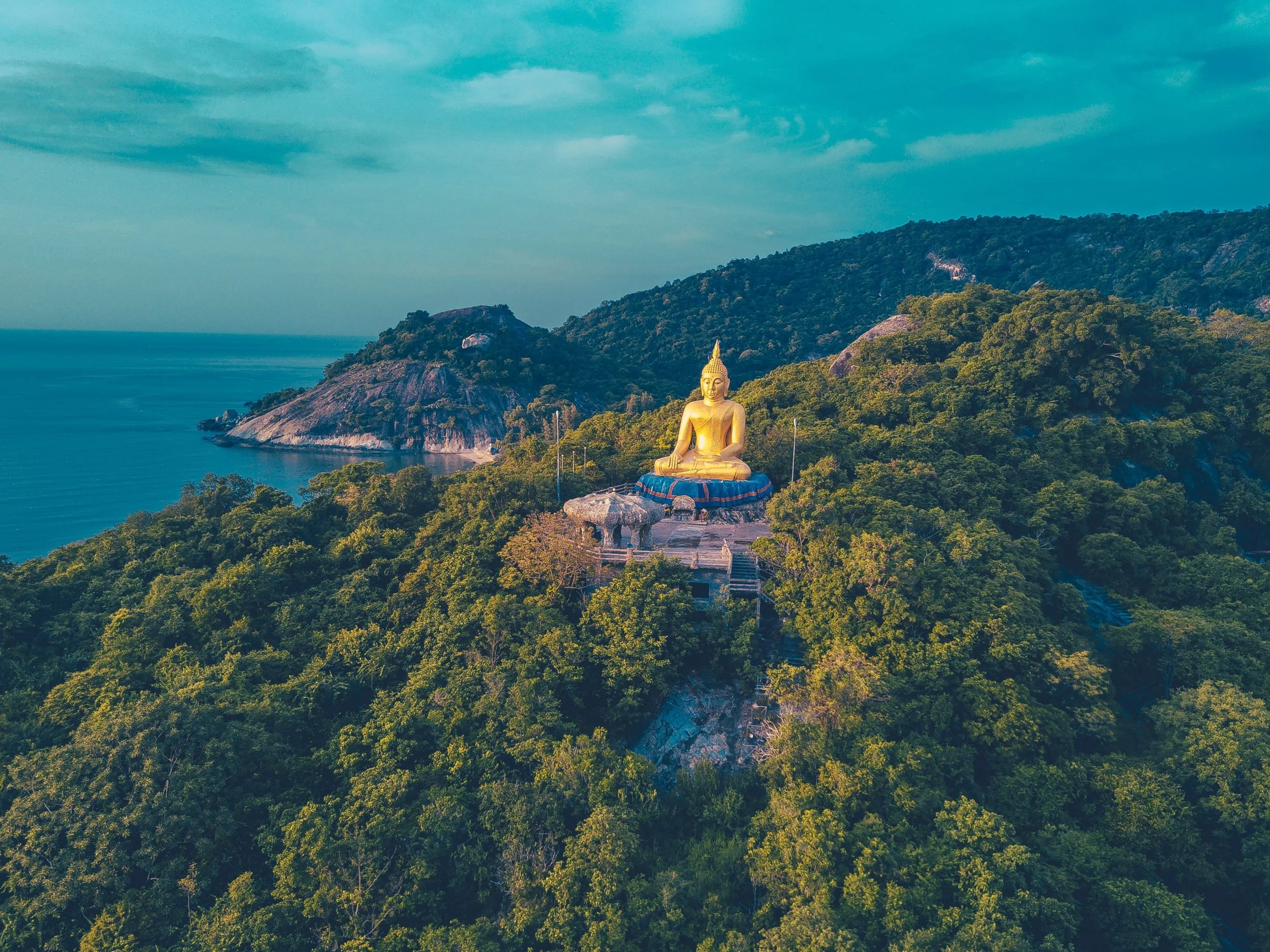 A large golden Buddha statue seated in meditation position outdoors on a blue platform, surrounded by lush green trees, with mountain hills and a partly cloudy sky in the background.