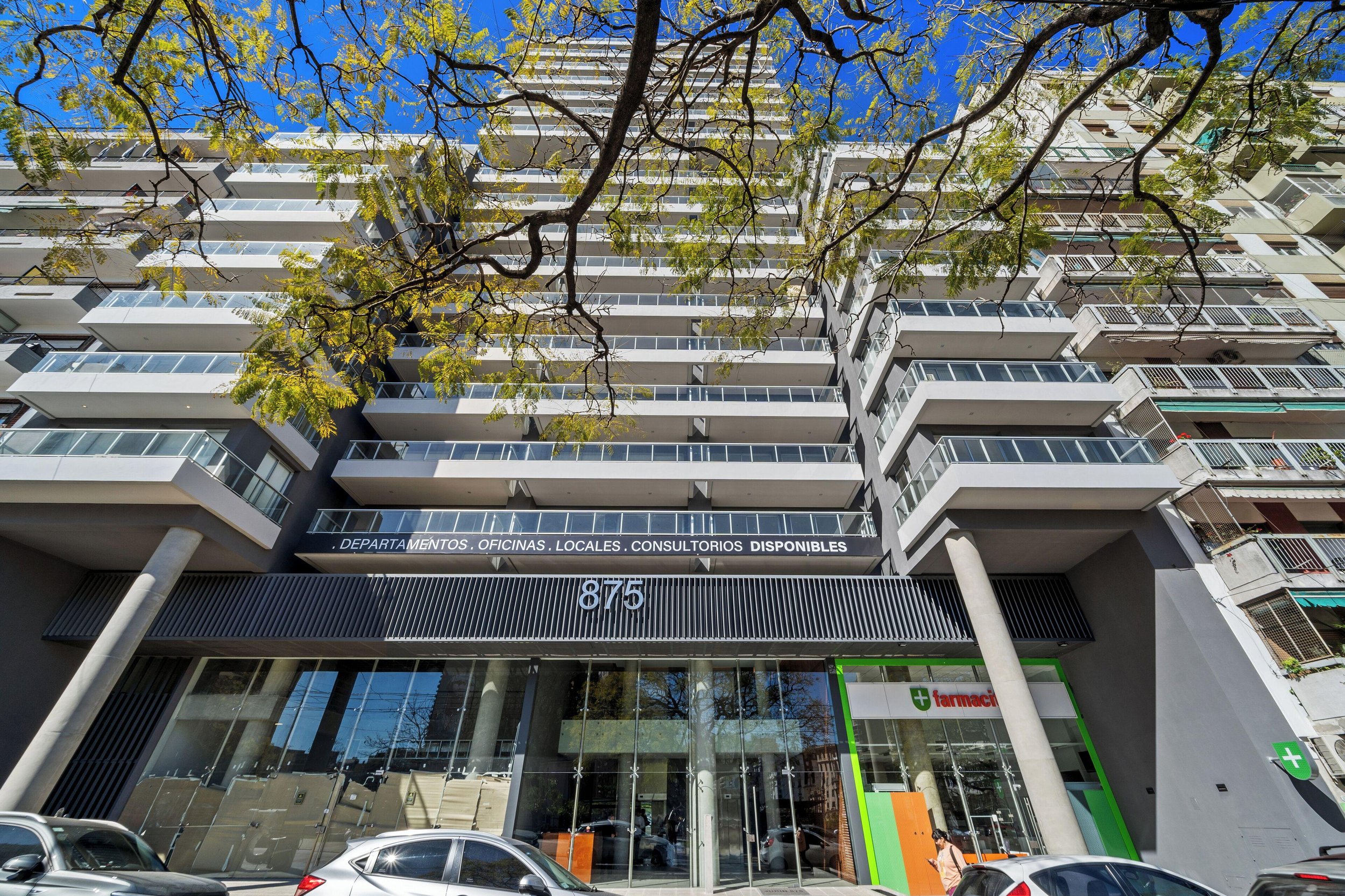Modern multi-story building with glass facade, balconies, and a commercial sign at the ground level, seen from street with parked cars and a tree with green leaves and a clear blue sky above.
