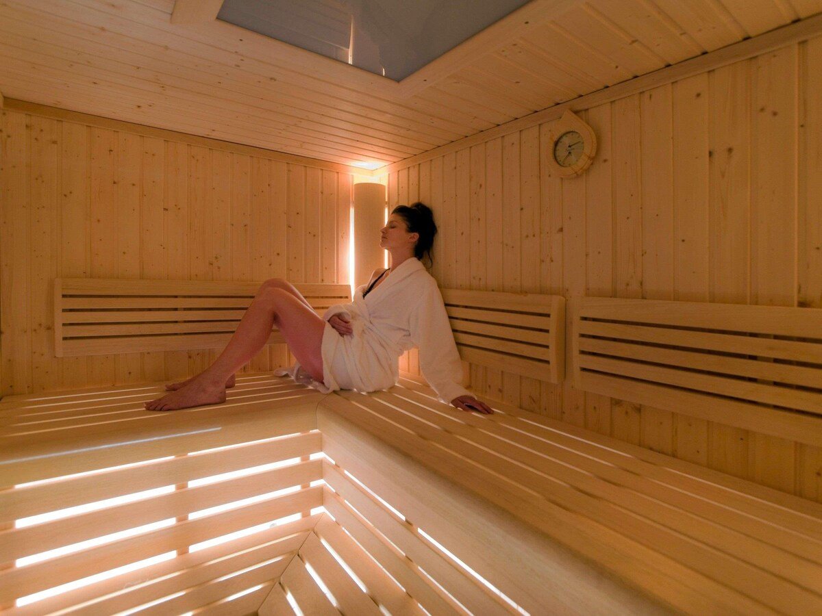 A woman in a white bathrobe relaxing inside a wooden sauna with light filtering through a small window.