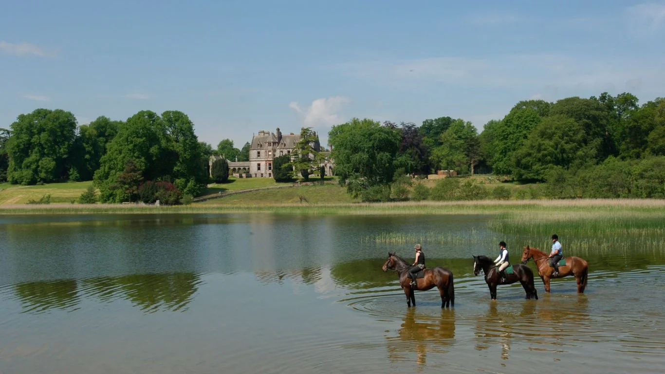 Three people riding horses through a lake near a large mansion surrounded by trees.