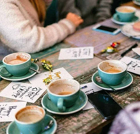 A woman in a long dress and straw hat stands overlooking a landscape of rock formations and numerous hot air balloons in the sky, while a man with a camera, wearing a hat and backpack, takes a picture of the scene during sunset or sunrise.
