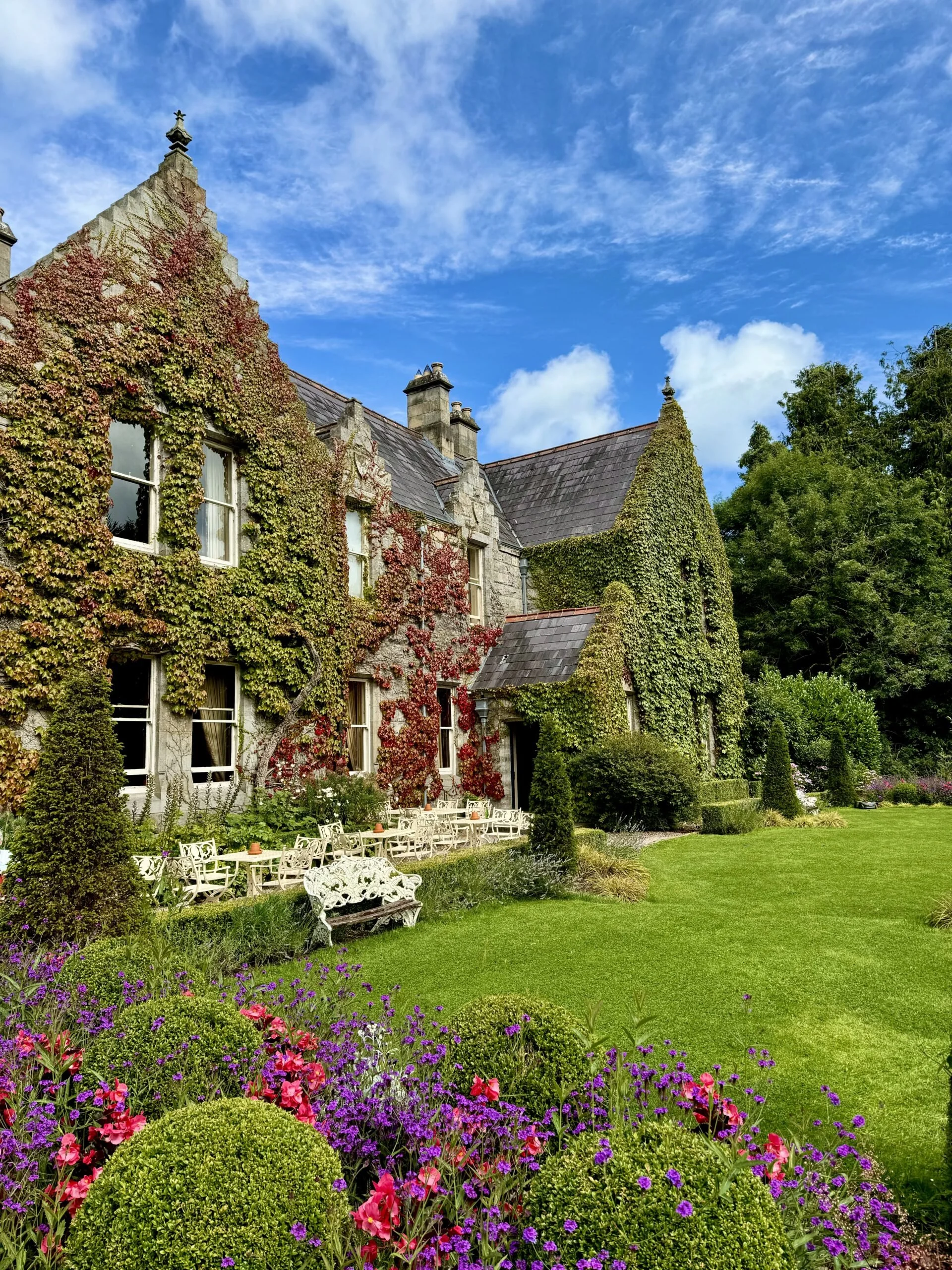 A stone house covered in green and red ivy, with a manicured lawn and colorful flower bed in the foreground, under a partly cloudy sky.