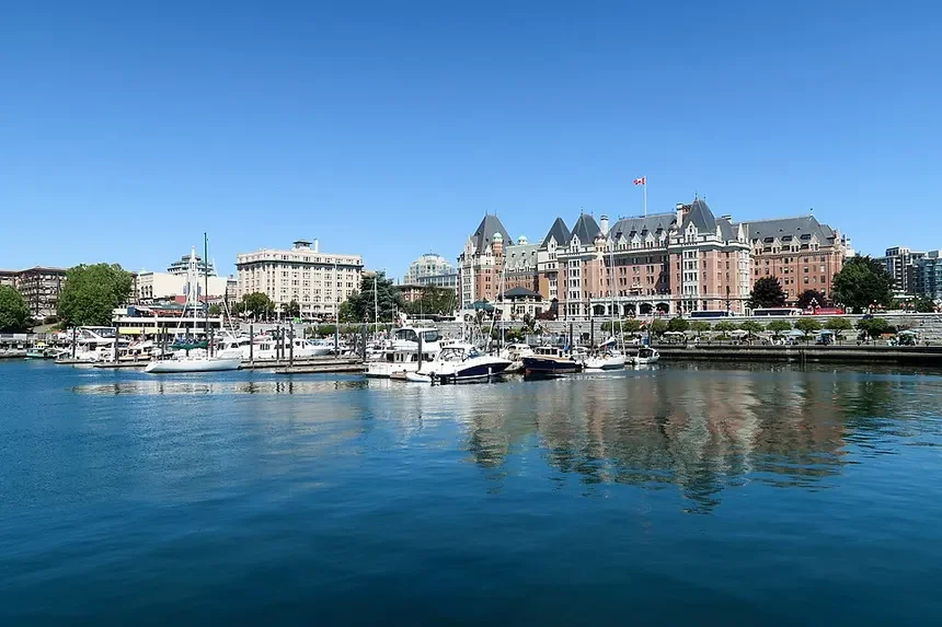 A marina with boats docked in the water, a historic-style building with turrets and a Canadian flag on top, and other city buildings in the background on a clear, sunny day.