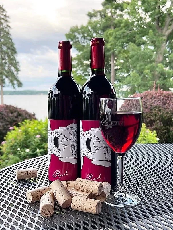 Two bottles of red wine with matching labels, a glass of red wine, and several wine corks on a metal outdoor table with a natural landscape background.