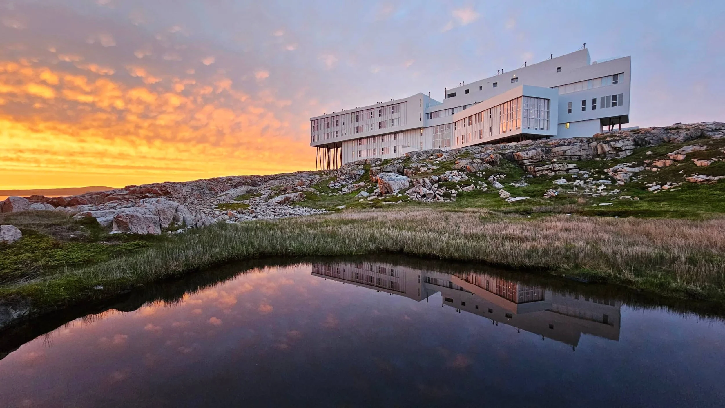 Modern white building on rocky hillside reflecting in a small pond during sunset with colorful sky and grassy landscape