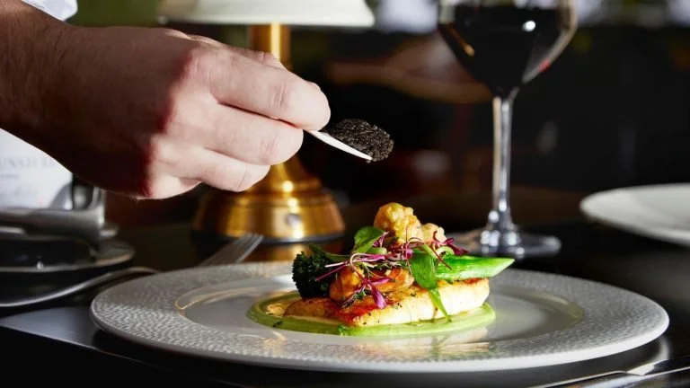 Person sprinkling black pepper on a plated gourmet dish with vegetables and sauce, with a glass of red wine in the background.