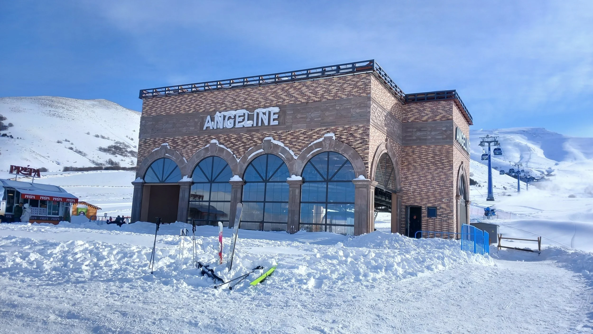 A snowy mountain resort with a large brick building labeled 'Angeline,' ski equipment in the snow, and ski lift chairs ascending the mountain in the background.