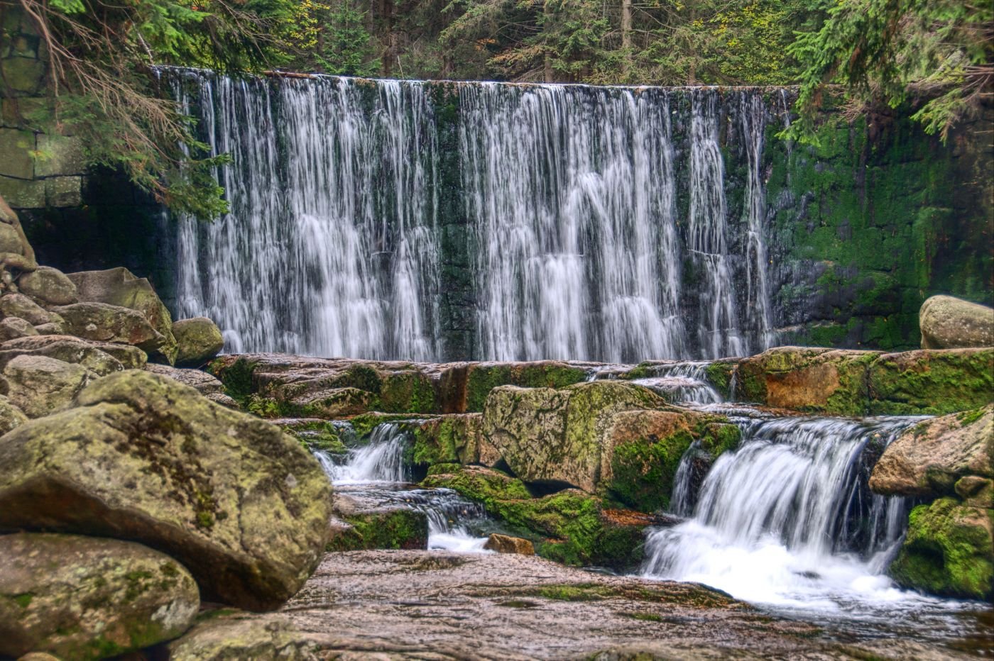 A waterfall flowing over a moss-covered rocky ledge in a forest with green trees in the background.