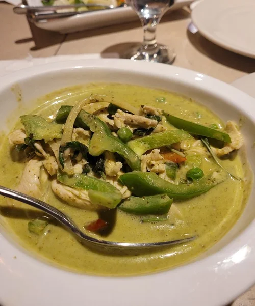 Bowl of green curry with vegetables including zucchini, peas, and chicken, on a table with a spoon in the curry, a glass of water, and a plate in the background.