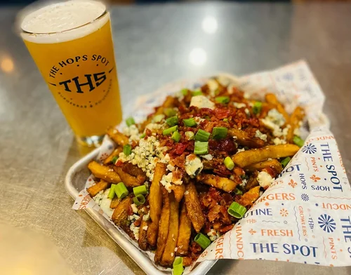 A tray of loaded fries topped with green onions, blue cheese crumbles, bacon bits, and ground meat served with a pint of beer in a glass labeled "The Hops Spot."