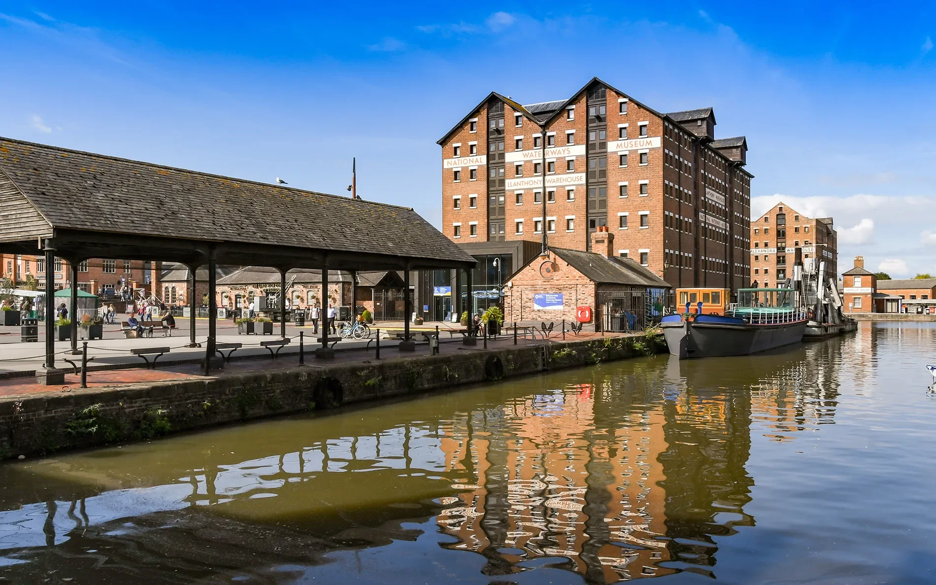 View of a historic waterfront area with a canal, a warehouse building labeled 'National Waterways Museum Llanhydny Warehouse,' a docked boat, and people walking along the promenade under a clear blue sky.