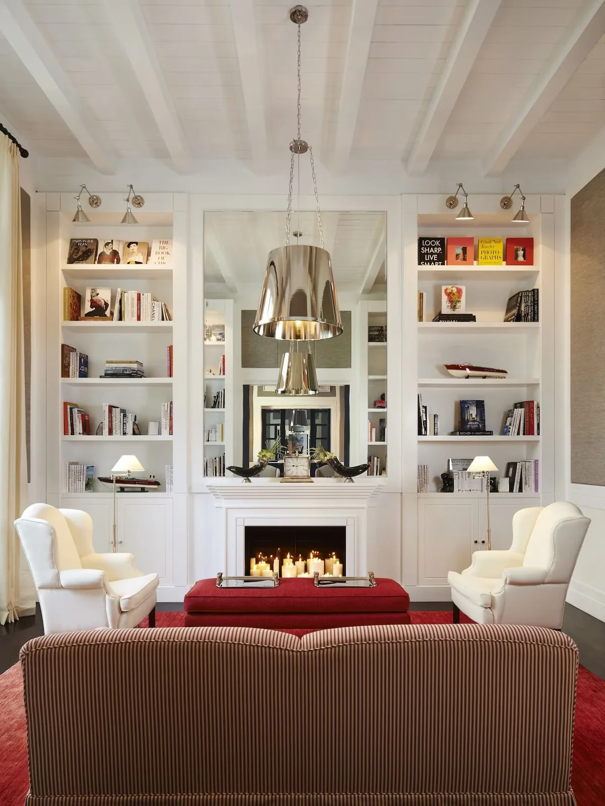 Living room with white built-in bookshelves, fireplace, mirror, two white armchairs, red ottoman with candles, and metallic pendant lights.