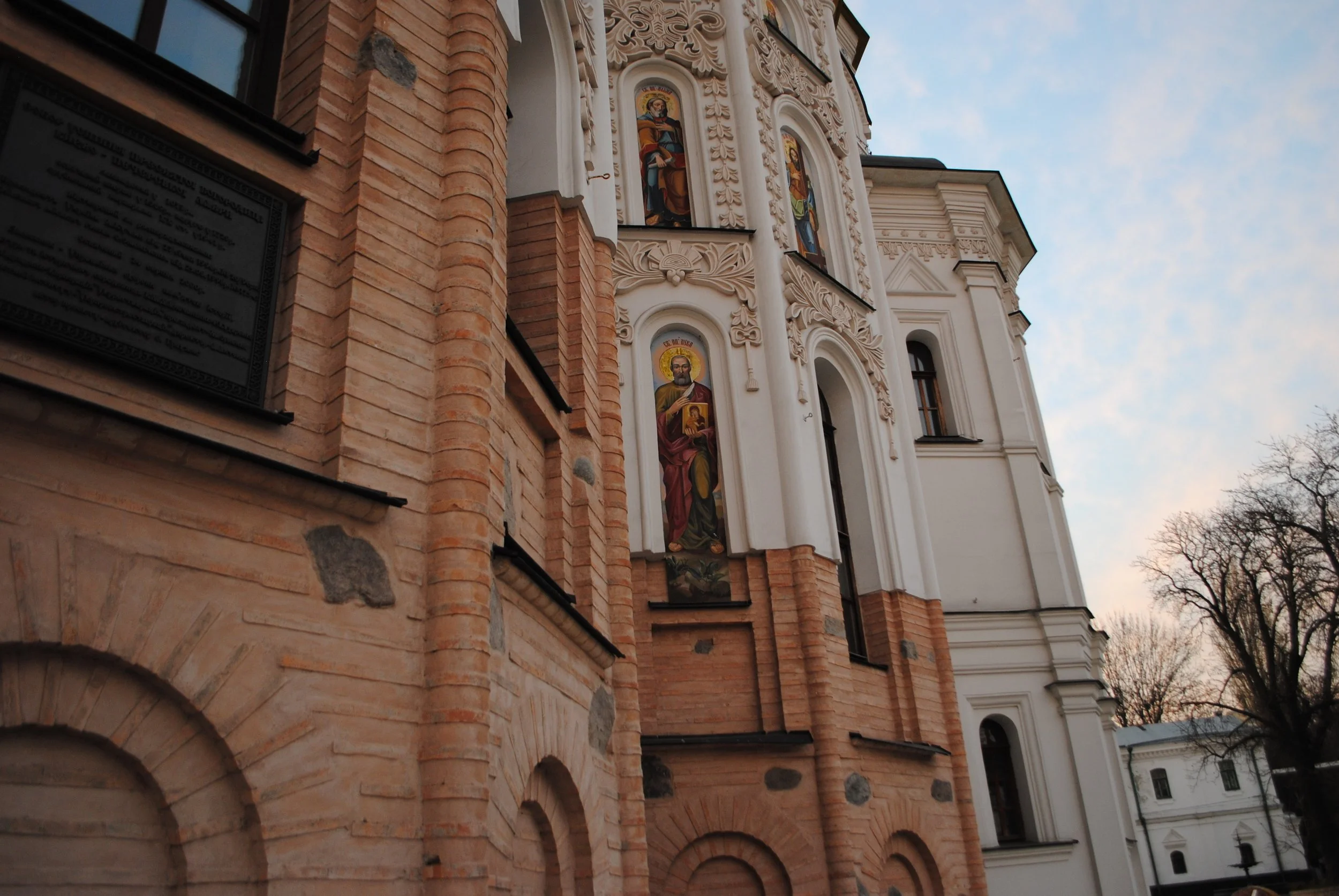The exterior of a church with ornate stucco decorations and biblical painted icons on the white facade, contrasted with red brickwork and arched windows, under a partly cloudy sky.