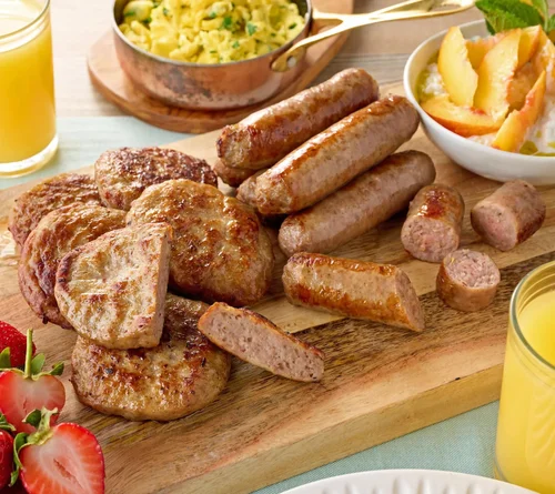 A wooden board with cooked breakfast meats including sausages and sausage patties, sides of scrambled eggs with herbs, sliced fried potatoes, and fresh strawberries, with glasses of orange juice.