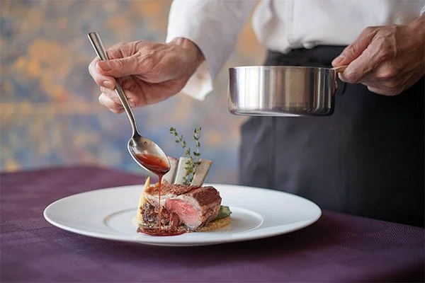Chef drizzling sauce over a plate with a cooked steak on it.
